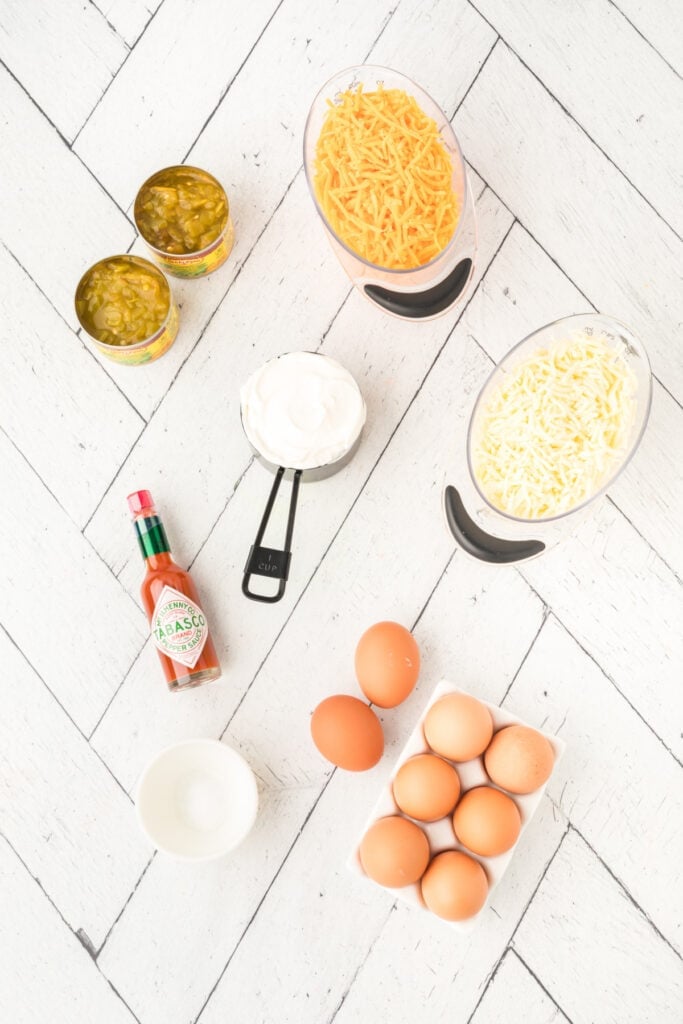 Ingredients for Chilies Rellenos Bake including eggs, shredded cheese, sour cream, green chilies, and hot sauce on a white surface
