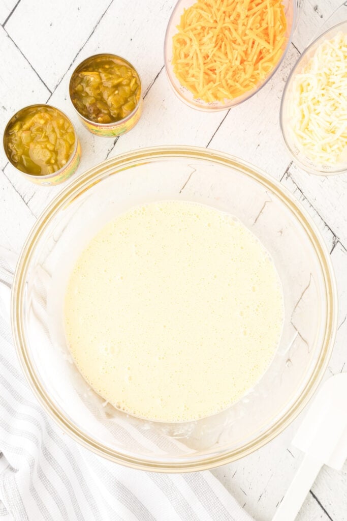 Whisked egg mixture in a glass bowl with green chilies and shredded cheese nearby