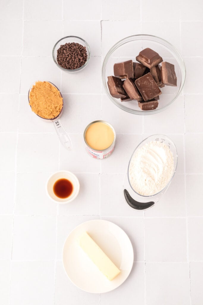 Overhead view of ingredients in bowls and plates for cookie dough truffles, including flour, butter, brown sugar, sweetened condensed milk, vanilla, mini chocolate chips, and chocolate candy coating.