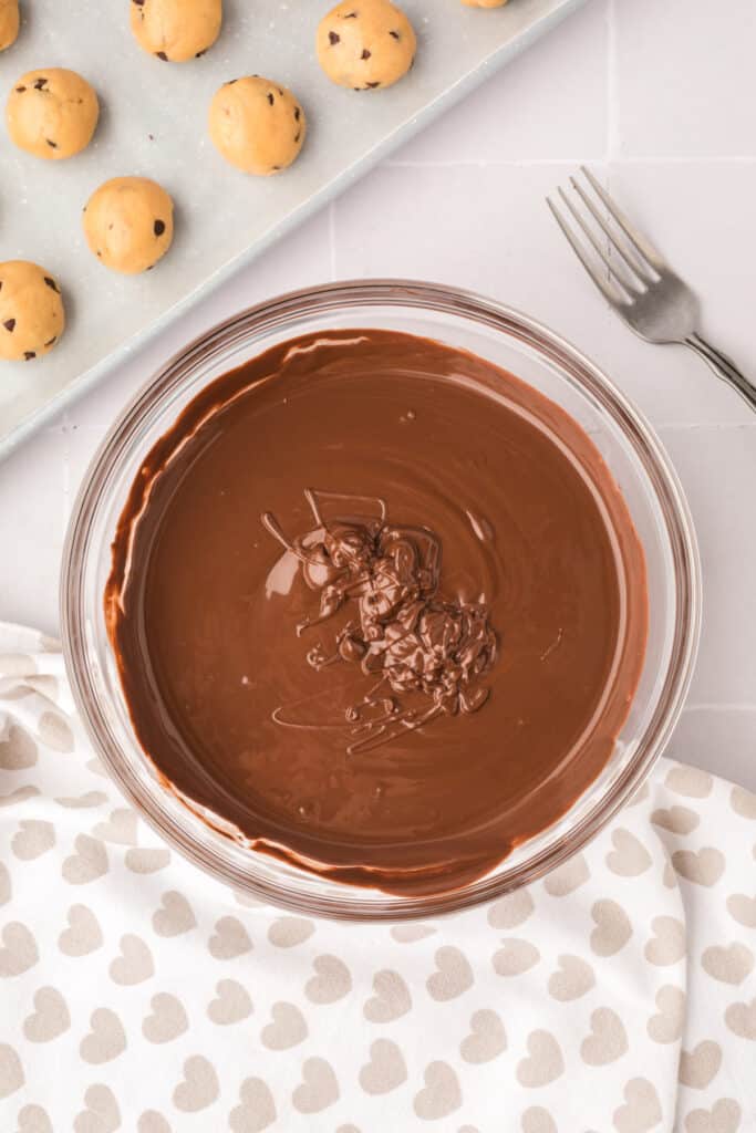 Bowl of melted chocolate with a fork beside it and cookie dough balls on a baking sheet ready for dipping.