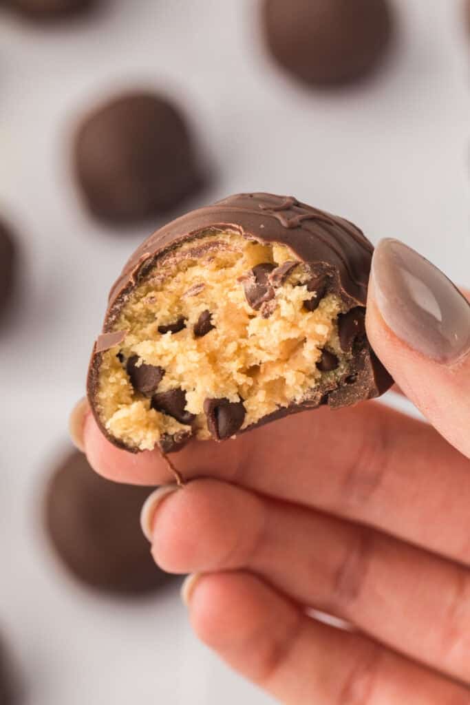 Hand holding a bitten chocolate truffle showing edible cookie dough center with mini chocolate chips.