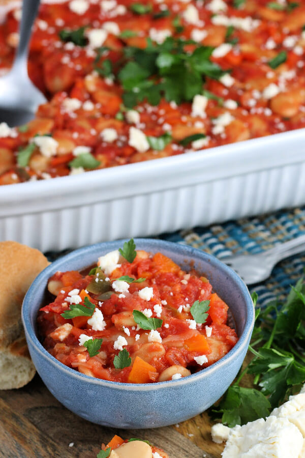 a serving of Greek Baked Beans in a blue bowl with the casserole in the background