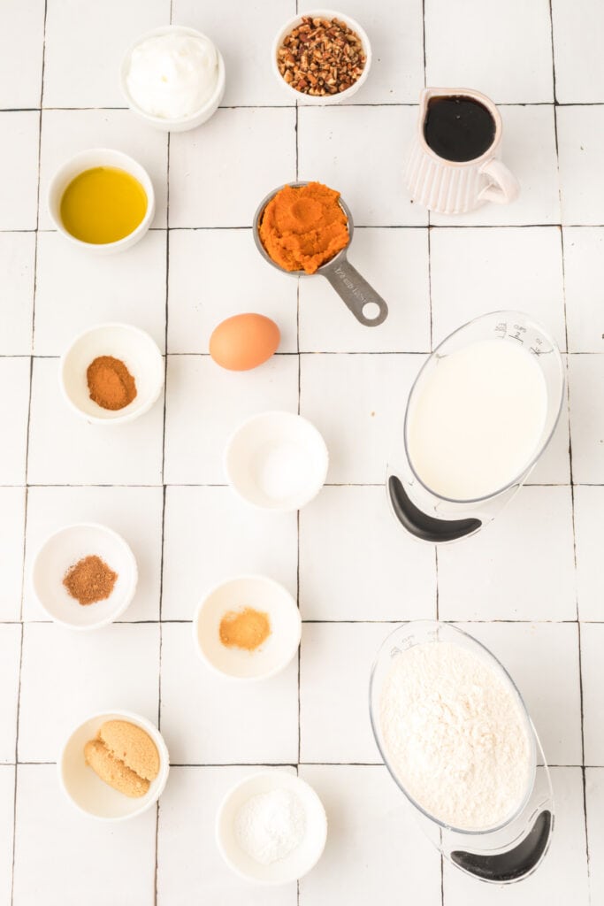 Overhead view of measured ingredients for pumpkin pancakes including pumpkin purée, milk, spices, flour, and egg on a white tiled surface