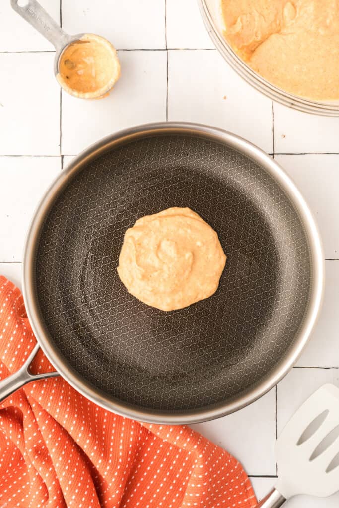 Pumpkin pancake cooking in frying pan with ladle and batter bowl in the background