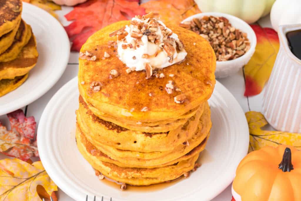 Overhead view of pumpkin pancakes on a white plate topped with whipped cream and pecans, surrounded by fall leaves