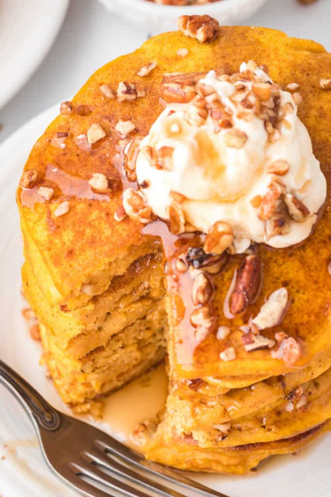 Stack of pumpkin pancakes with syrup and whipped cream, showing a cut section revealing soft, fluffy texture