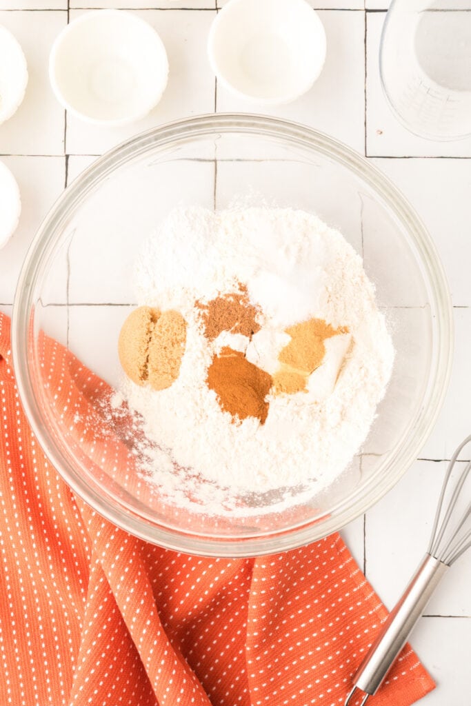 Mixing bowl filled with flour, brown sugar, and pumpkin spices on white tiled surface with orange kitchen towel