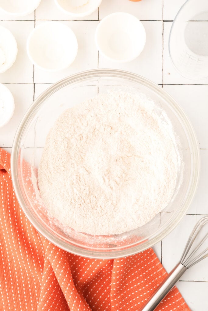 Bowl of combined dry ingredients for pumpkin pancakes with whisk and orange towel beside it