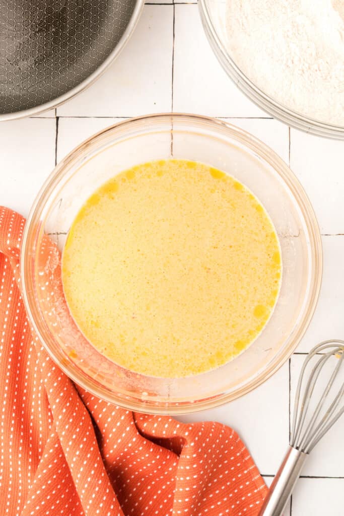 Whisked wet ingredients for pumpkin pancakes in a glass bowl beside a pan and orange towel