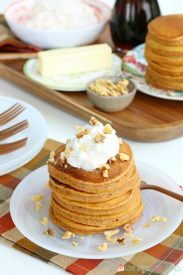 Pumpkin pancakes stacked up on a plate with nuts and whip cream on top.