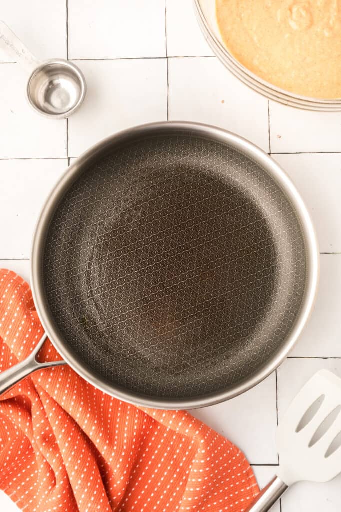 Oiled frying pan heating on stovetop beside pumpkin pancake batter and measuring cup