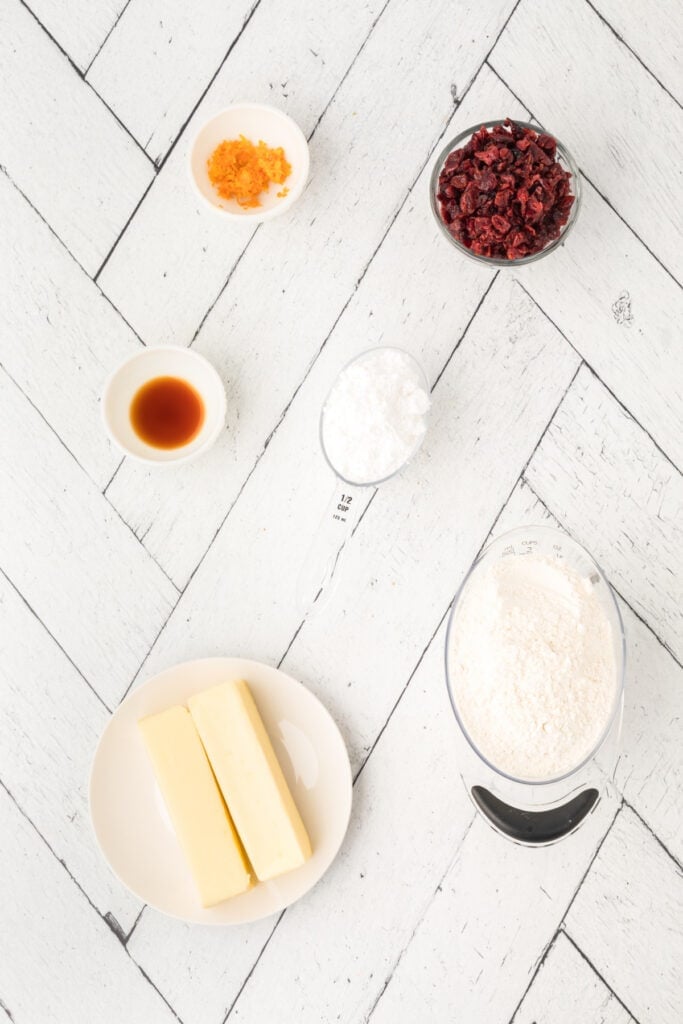 Overhead shot of ingredients for Cranberry Orange Christmas Sandies, including butter, flour, powdered sugar, vanilla, orange zest, and dried cranberries arranged on a white herringbone surface.