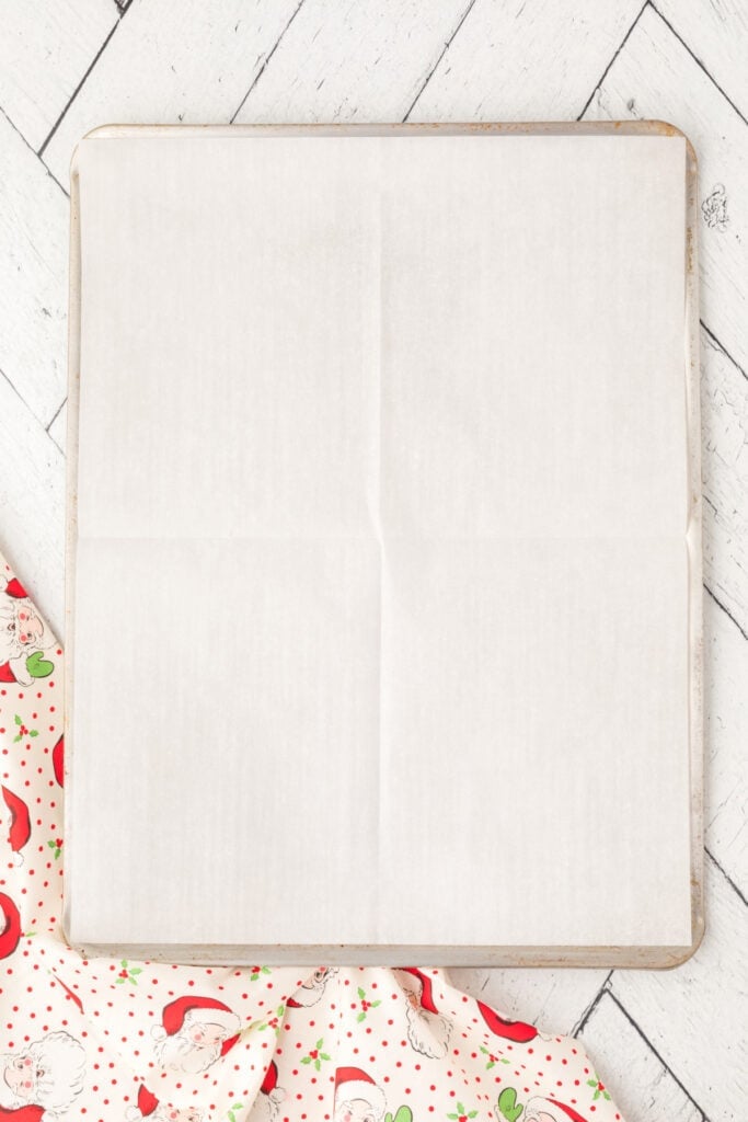 Baking sheet lined with parchment paper, ready for shaping Cranberry Orange Christmas Sandies dough, with festive Santa-patterned fabric underneath.