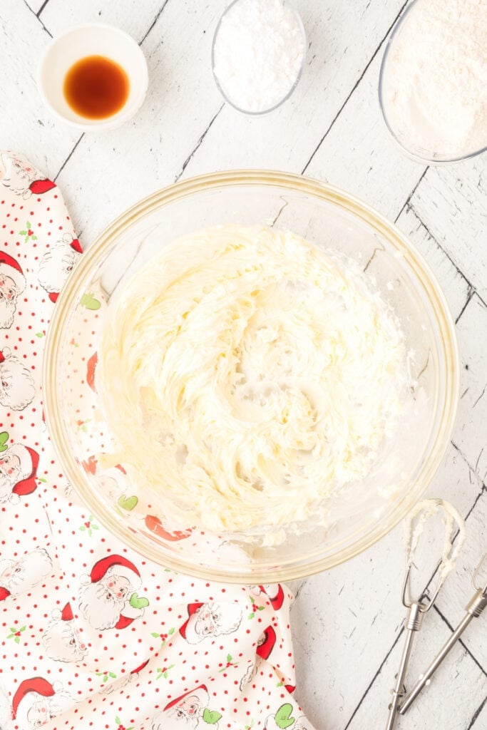 Butter creamed in a glass mixing bowl with electric beaters nearby, next to bowls of vanilla, powdered sugar, and flour on a white wood surface.