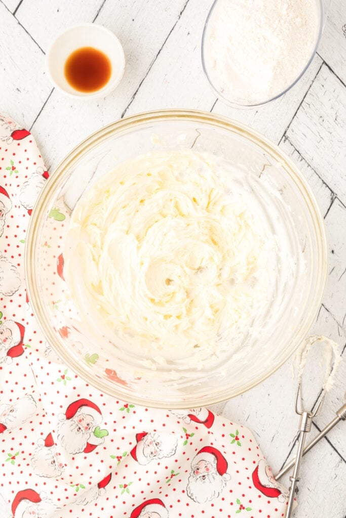 Smooth creamed butter and sugar mixture in a glass bowl with Santa fabric visible underneath, showing early stage of cookie dough mixing.