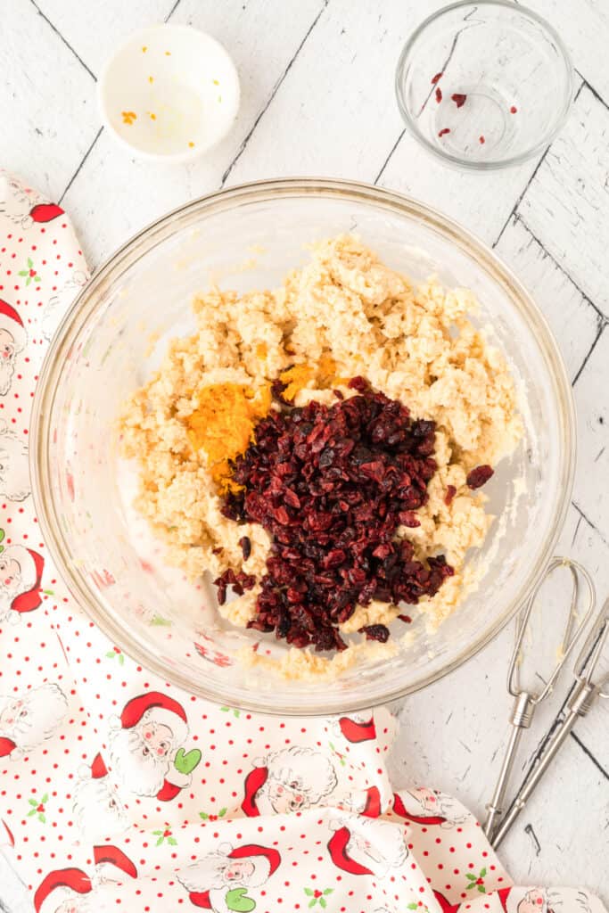 Bowl of cookie dough with dried cranberries and orange zest added on top, ready to mix for Cranberry Orange Christmas Sandies.