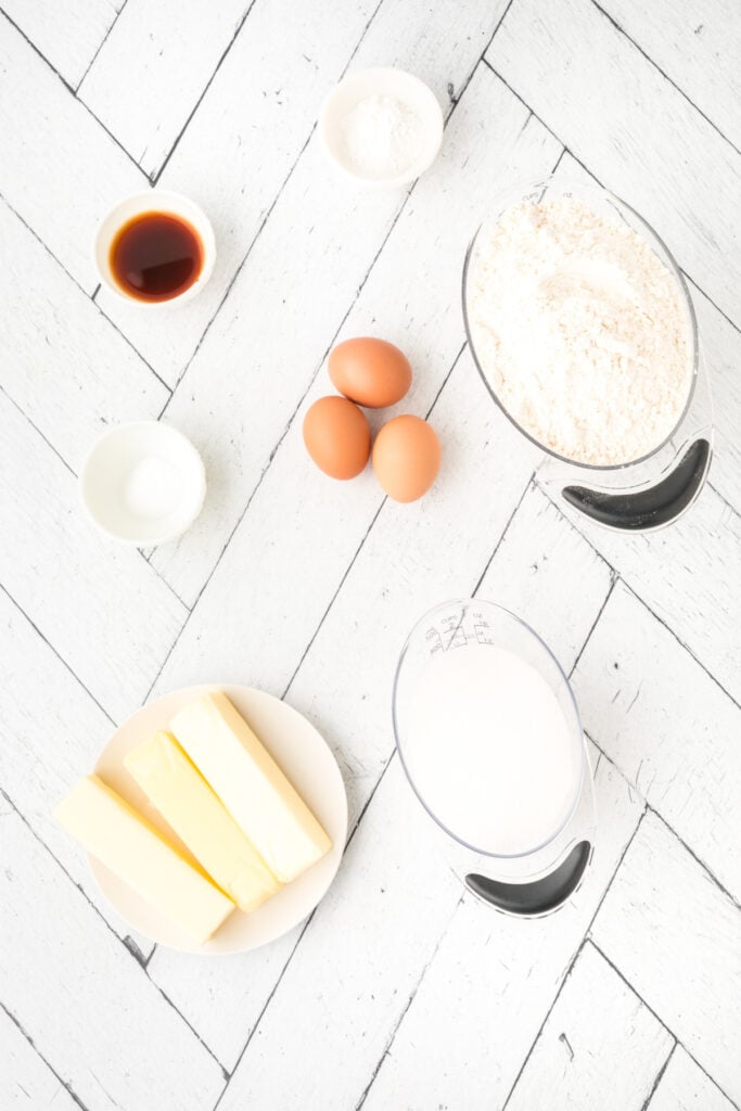 Overhead view of measured ingredients for soft sugar cookies including butter, eggs, flour, sugar, vanilla, salt, and baking powder on a white wood surface.
