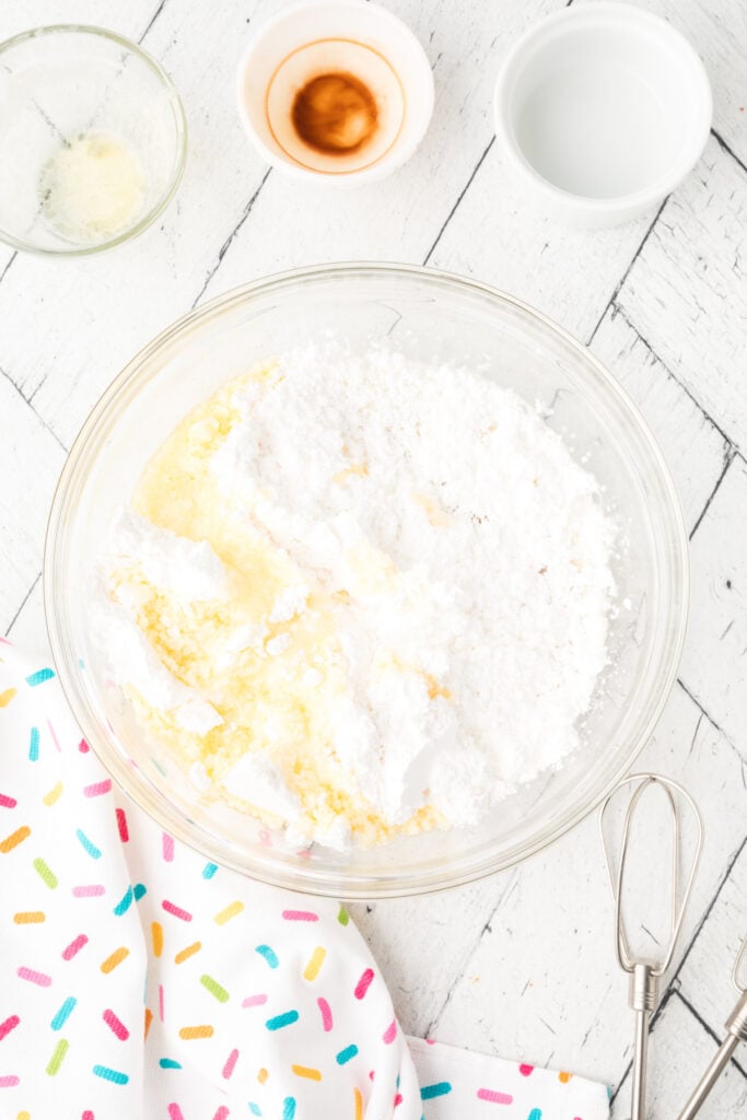 Frosting ingredients including powdered sugar, melted butter, milk, vanilla, food coloring, and sprinkles on a white background.