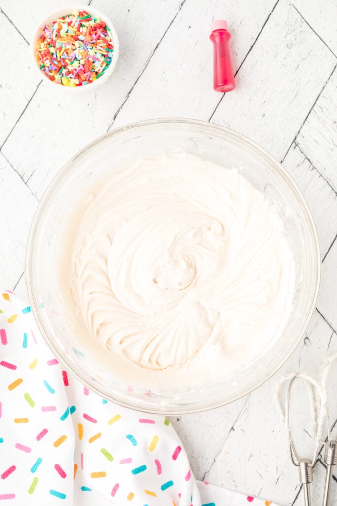 Bowl of fluffy white frosting with rainbow sprinkles and red food coloring bottle nearby.