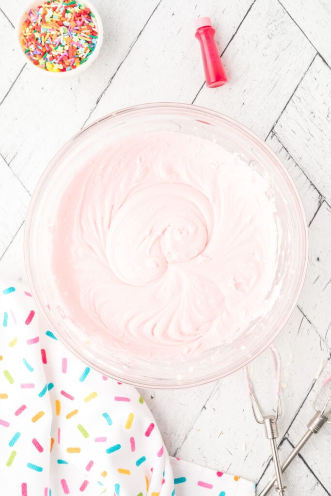 Bowl of light pink frosting after mixing in red food coloring, with sprinkles in background.