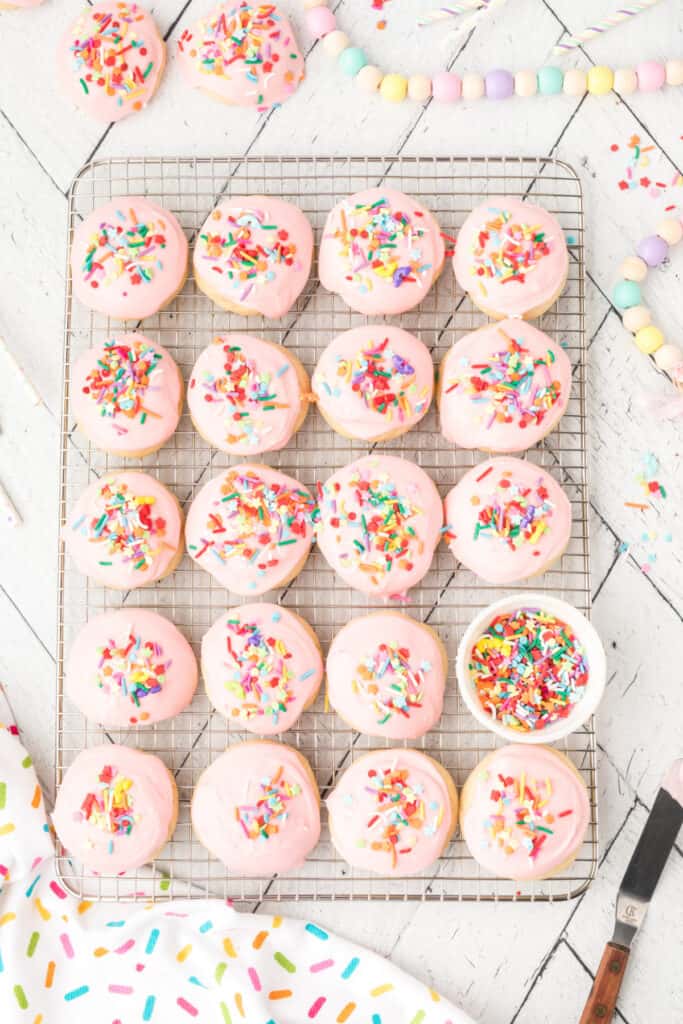 Frosted pink cookies topped with colorful rainbow sprinkles on a cooling rack.