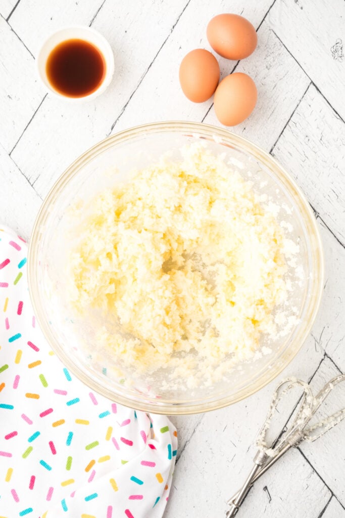 Butter and sugar creamed together in a mixing bowl with eggs and vanilla nearby on a white wood background.