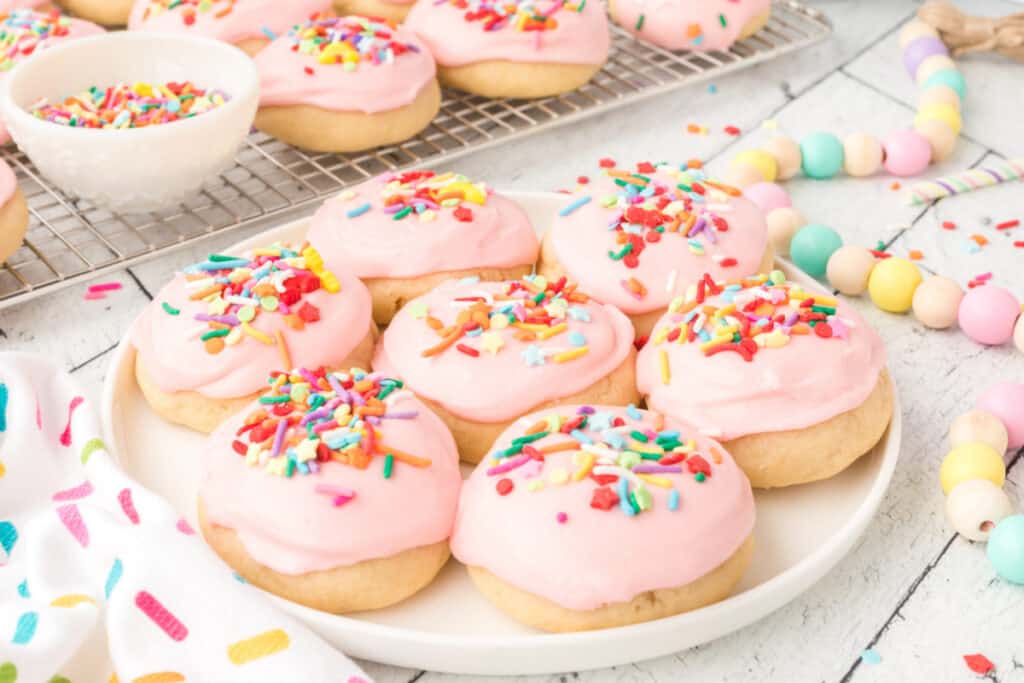Plate of frosted sugar cookies with pink icing and colorful sprinkles, pastel beads in background.
