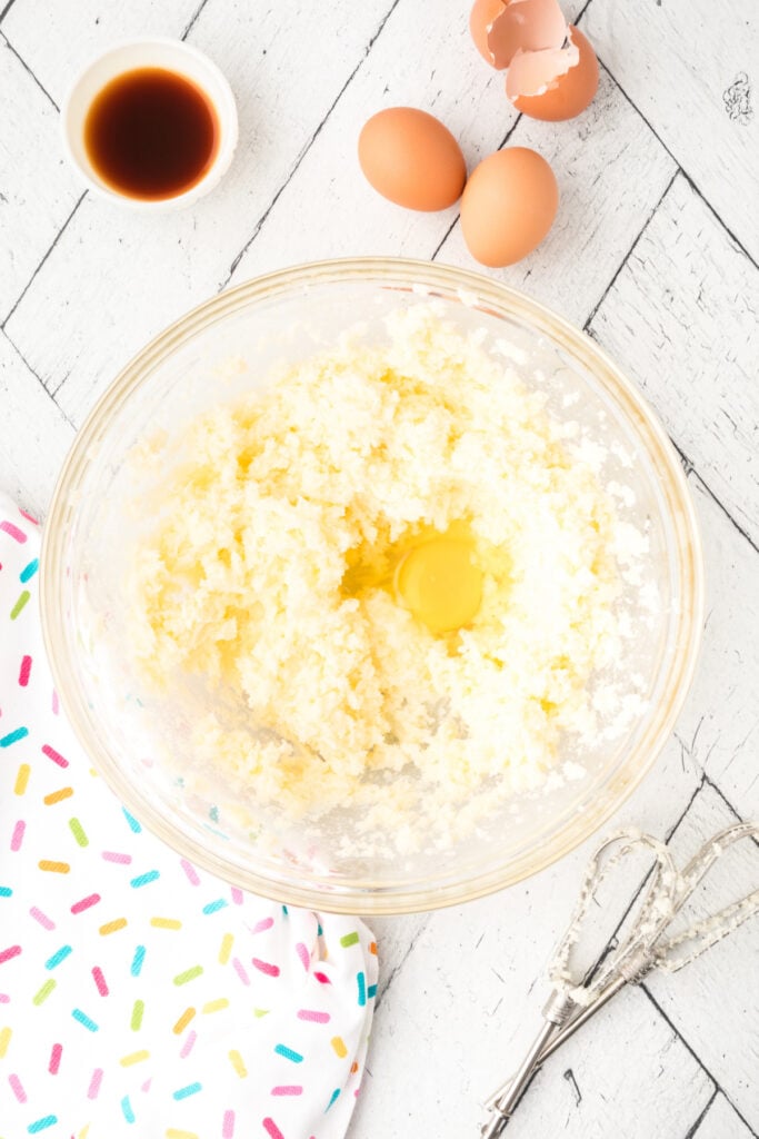 Egg added to the creamed butter and sugar mixture in a clear glass bowl with other eggs nearby.