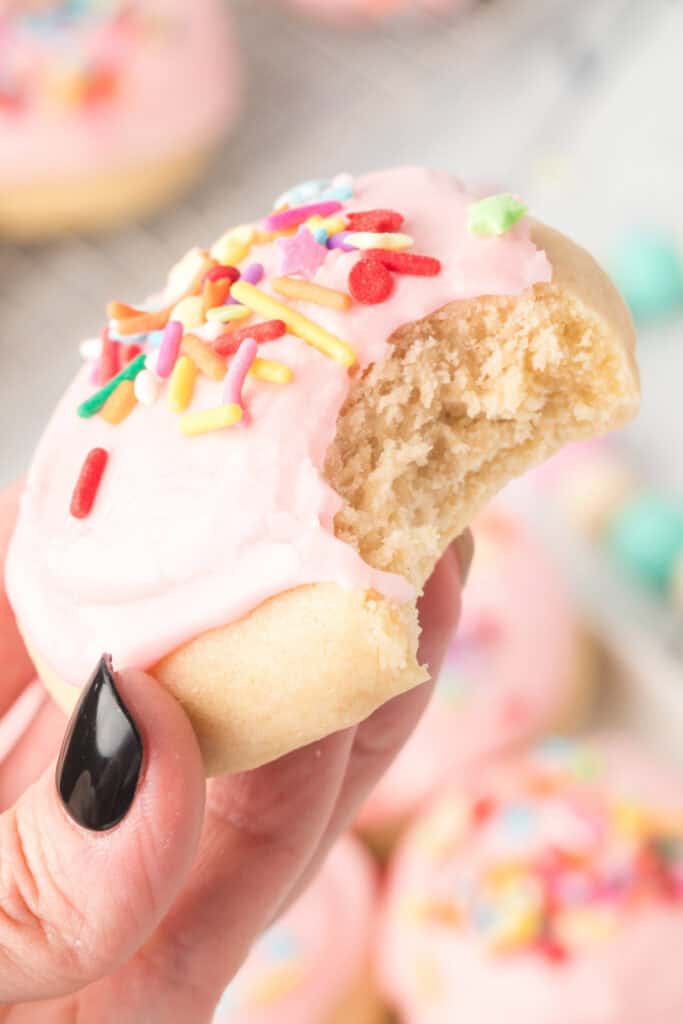 Soft sugar cookie with pink frosting and rainbow sprinkles held close up with bite missing.