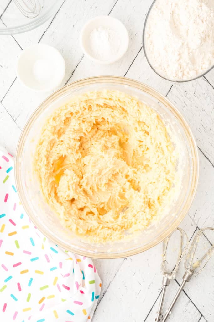 Soft sugar cookie dough in a glass mixing bowl with flour, baking powder, and salt in bowls nearby.