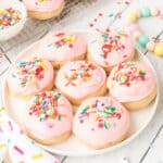 Square crop of soft sugar cookies with pink frosting and rainbow sprinkles on a plate.