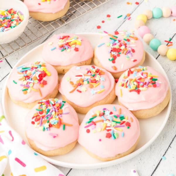 Square crop of soft sugar cookies with pink frosting and rainbow sprinkles on a plate.