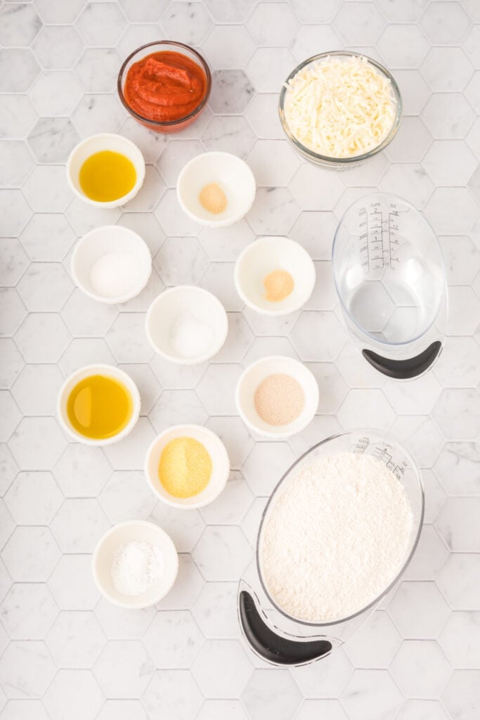 Ingredients for Bread Machine Pizza Dough arranged on a white countertop with flour, yeast, olive oil, pizza sauce, mozzarella cheese, and seasonings