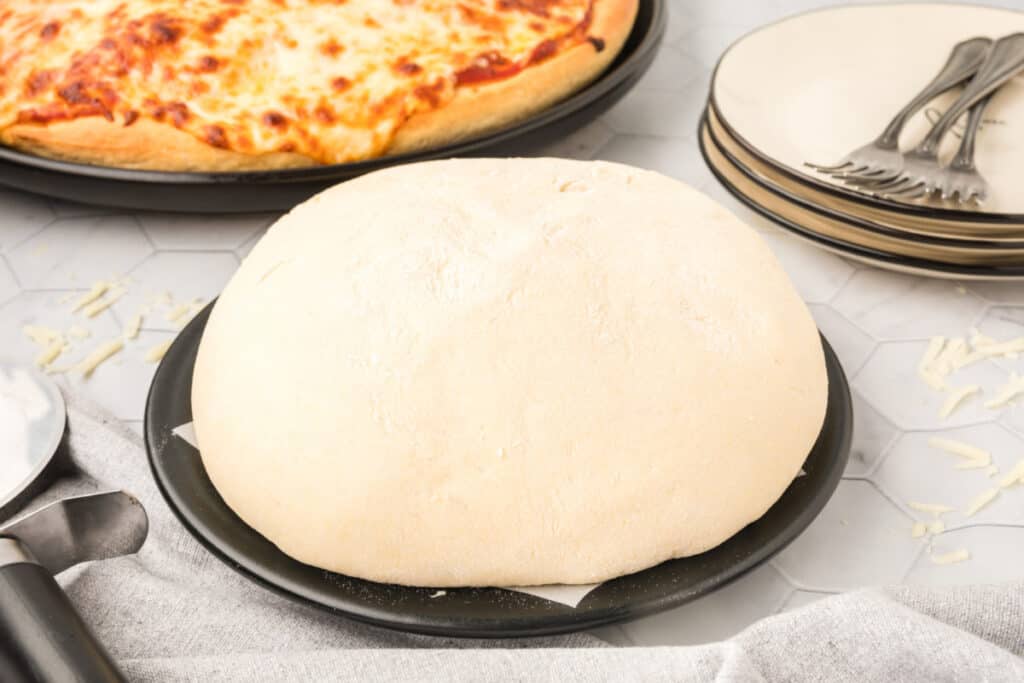 Homemade Bread Machine Pizza Dough shaped into a smooth pizza dough ball on a pizza pan with baked cheese pizza in the background