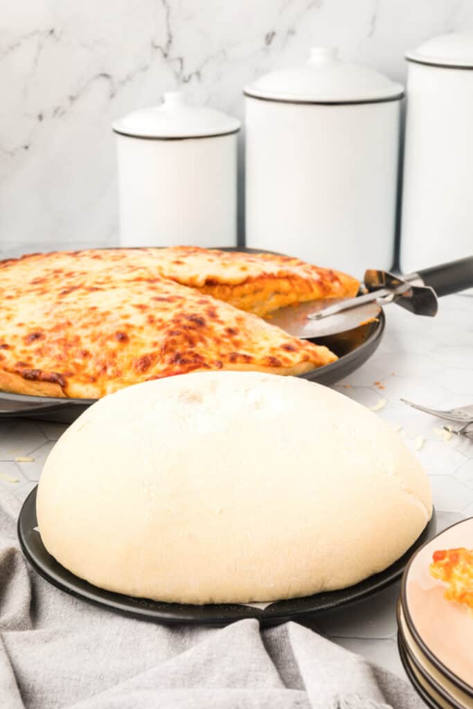 Prepared homemade Bread Machine Pizza Dough on a pizza pan beside a baked cheese pizza on a kitchen counter