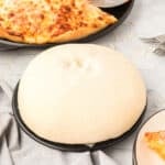 Square image of homemade Bread Machine Pizza Dough on a pizza pan with baked cheese pizza in the background