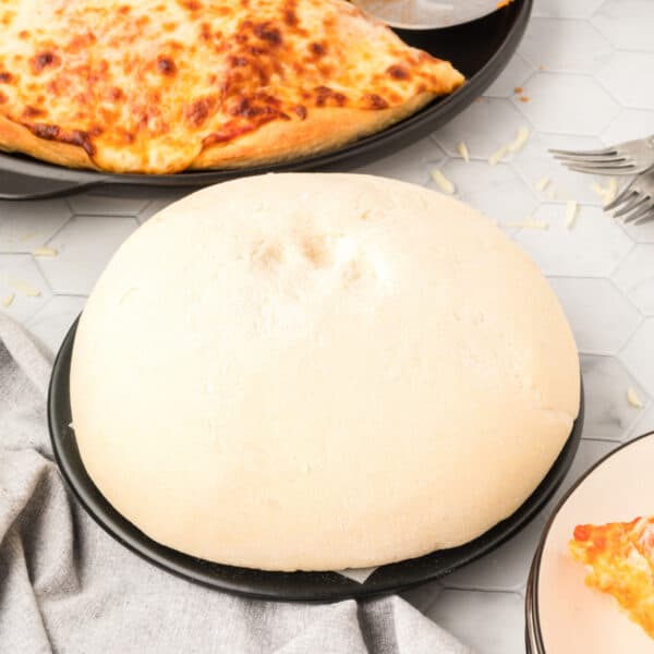 Square image of homemade Bread Machine Pizza Dough on a pizza pan with baked cheese pizza in the background