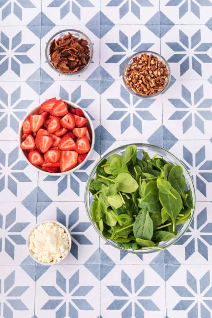 bowls of fresh spinach leaves, sliced strawberries, crumbled feta cheese, cooked bacon pieces, and chopped pecans on a patterned surface for strawberry spinach salad
