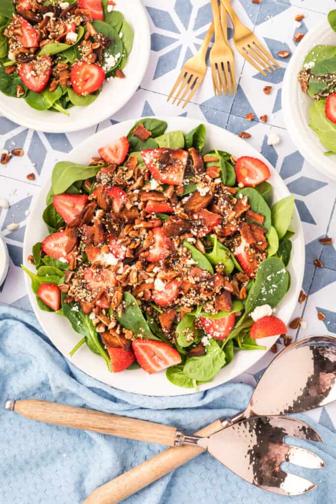 large bowl of strawberry spinach salad with balsamic poppy seed dressing, bacon, feta, pecans, and sliced strawberries with serving utensils