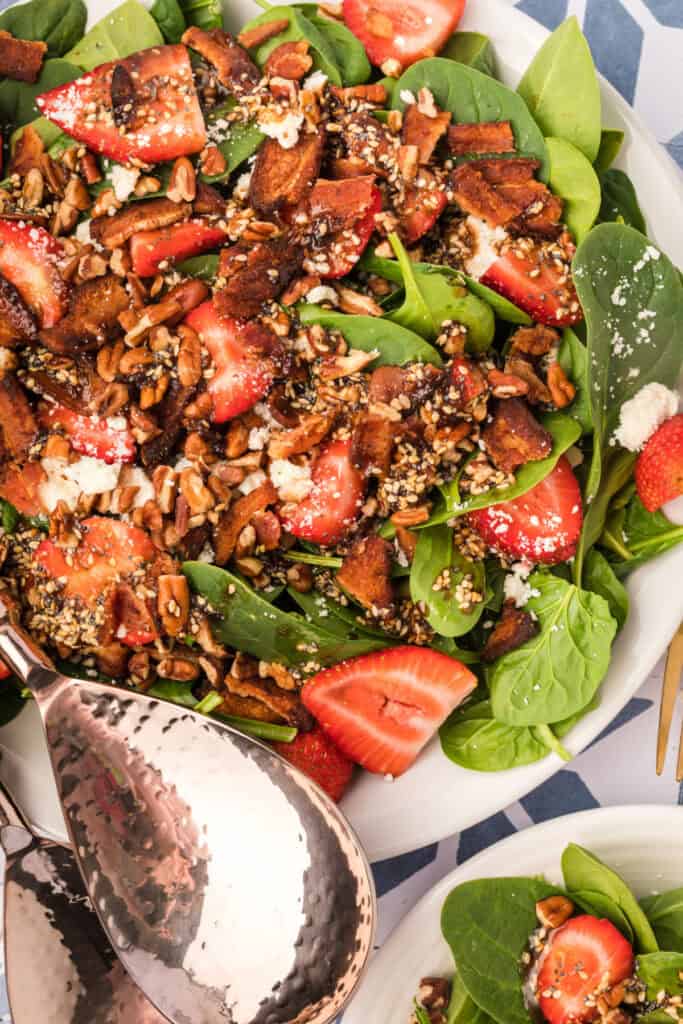 close-up of strawberry spinach salad with balsamic poppy seed dressing, bacon, feta, pecans, and sliced strawberries with serving utensils
