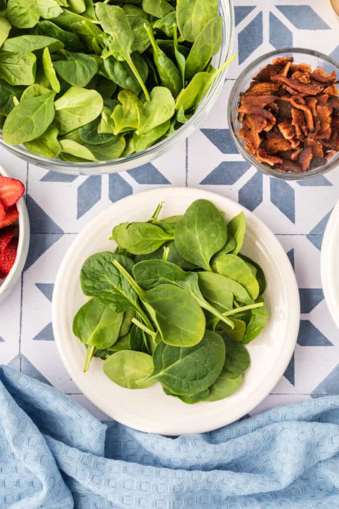plate of fresh spinach leaves with bowls of bacon and strawberries nearby for strawberry spinach salad preparation