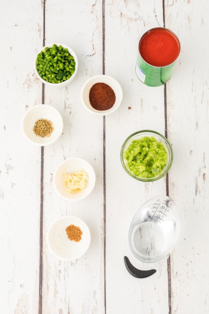 small bowls of chopped green bell pepper, jalapeno, garlic, spices, tomato sauce, and a measuring cup of water on a white wood surface