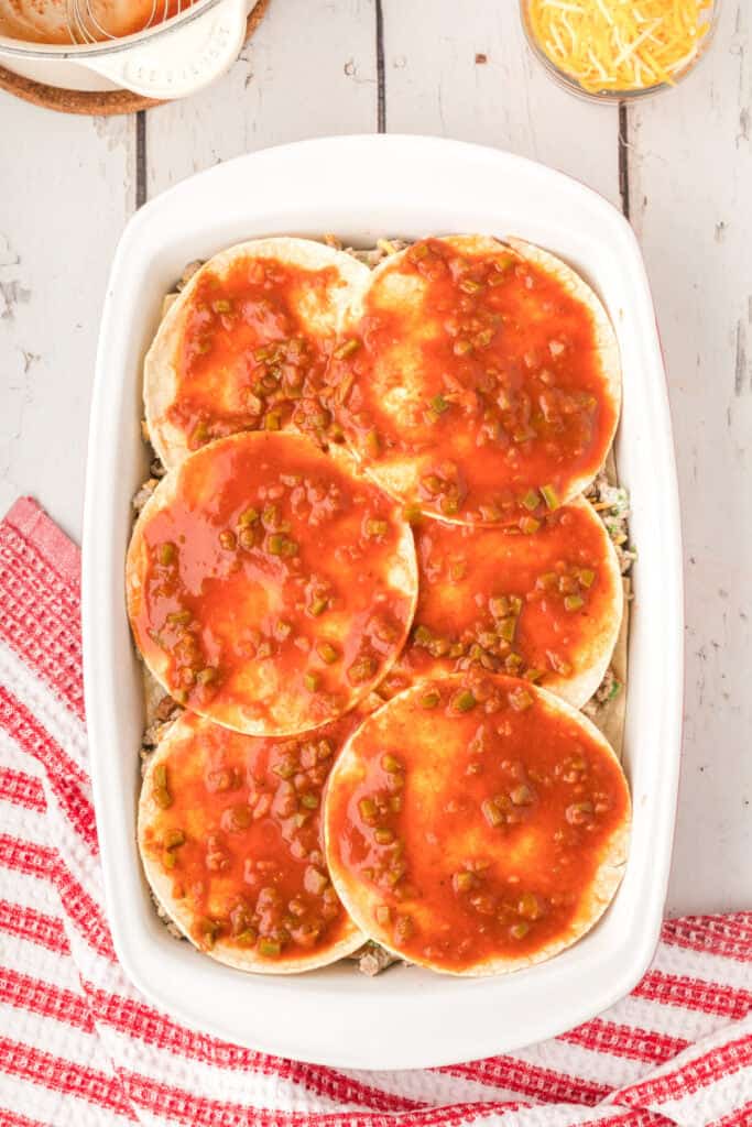 tortillas topped with enchilada sauce layered over beef mixture in a baking dish