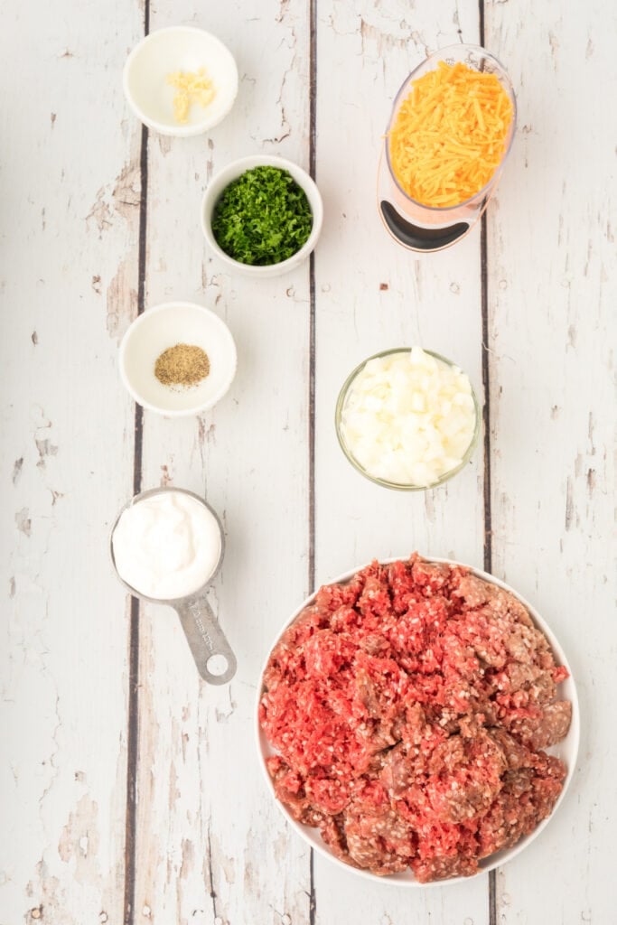 ground beef, chopped onion, shredded cheddar cheese, sour cream, parsley, garlic, and black pepper arranged in bowls on a white wood surface