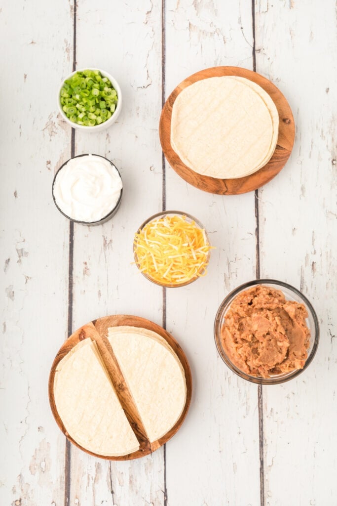 corn tortillas, refried beans, shredded cheese, sour cream, and sliced green onions on a white wood surface