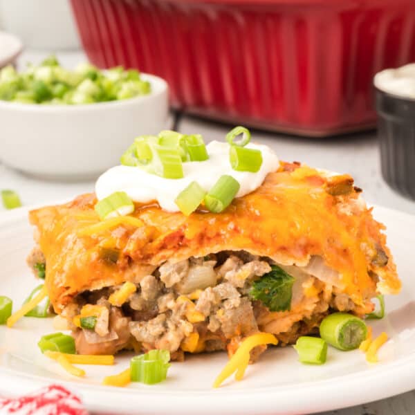 square image of beef and bean enchilada casserole slice topped with sour cream and green onions on a plate
