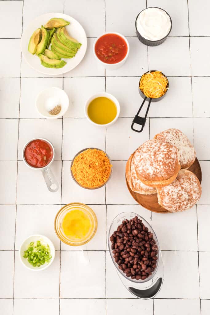 ingredients for Black Bean Nacho Burgers arranged on a white tile surface including black beans, buns, avocado, salsa, cheese, and oil