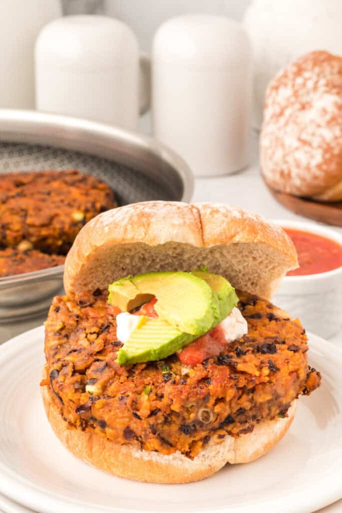 Black Bean Nacho Burger on a bun topped with avocado, sour cream, and salsa with cooked patties in the background