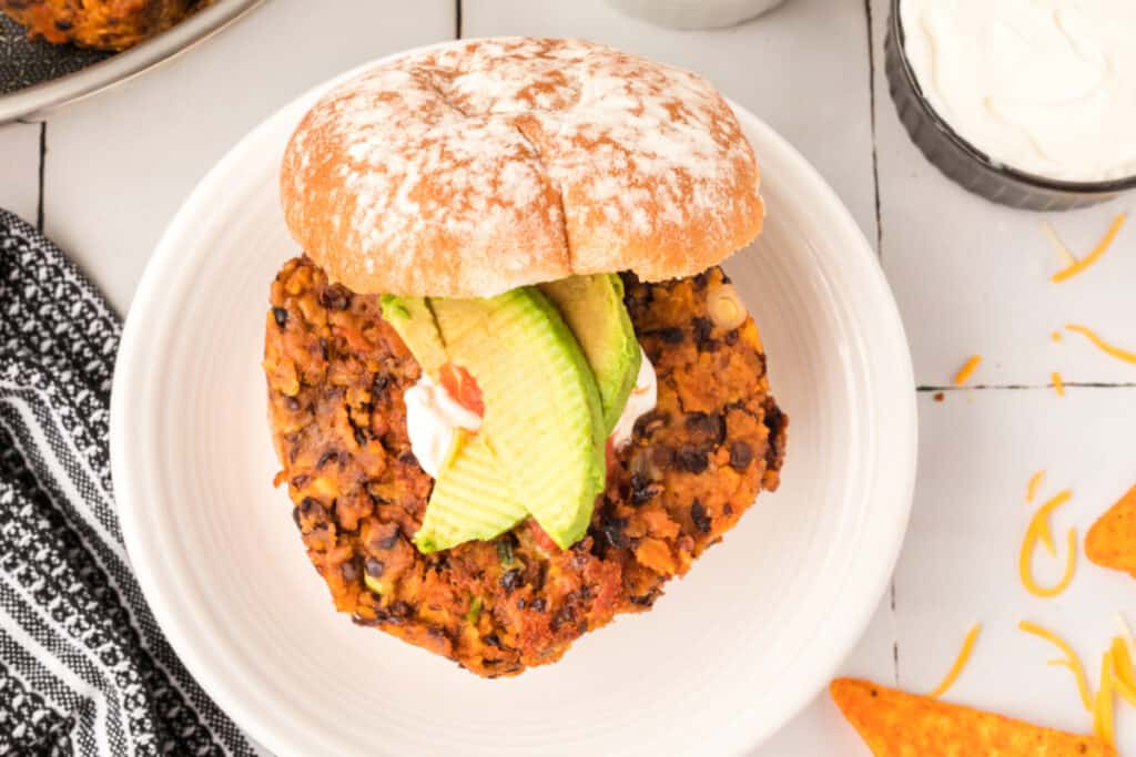 overhead view of Black Bean Nacho Burger on a bun with avocado, sour cream, and salsa on a white plate