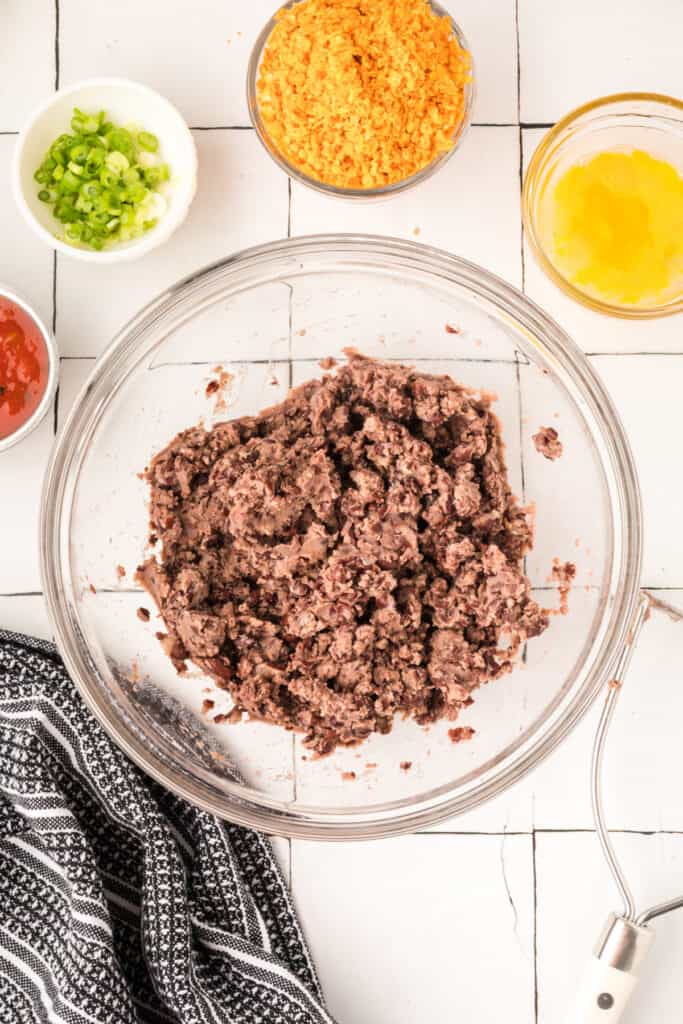 mashed black beans in a glass mixing bowl with chopped green onions, crushed chips, and egg nearby